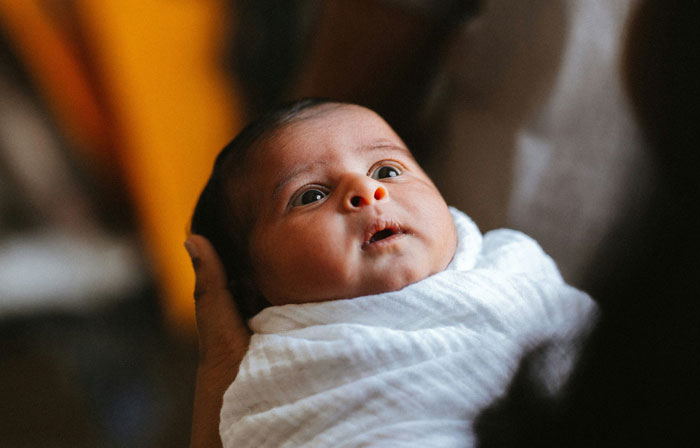 Baby wrapped in a white blanket, looking up with wide eyes, being held gently in someone's arms. Baby wrapped in a white blanket, looking up with wide eyes, being held gently in someone's arms.