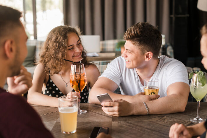 Young guys and a married woman smiling and drinking cocktails together at a bar table in a casual setting