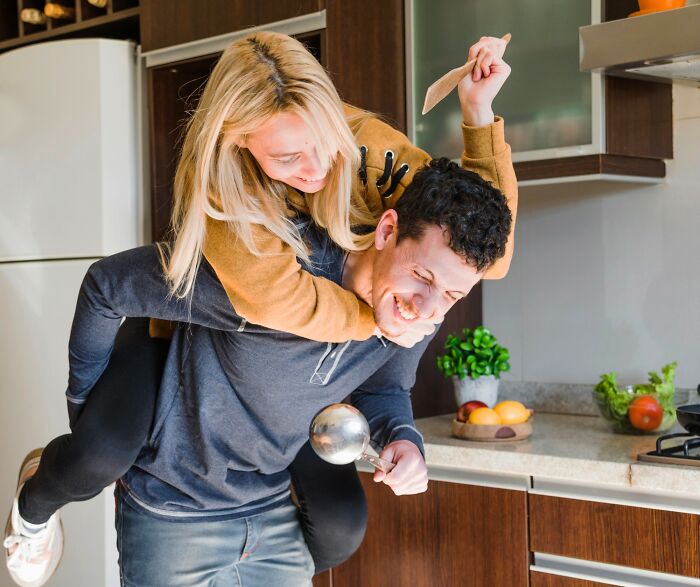 Woman playfully rides piggyback on man in kitchen, both holding utensils, highlighting dating dynamics.