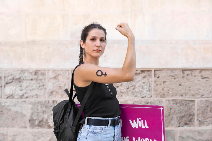 Woman confidently flexing arm with symbol tattoo, challenging common assumptions, holding a pink protest sign.