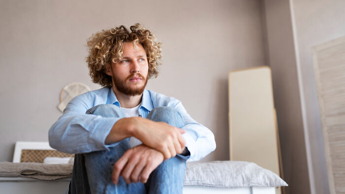 Young man with curly hair sitting on bed, looking thoughtful and contemplative about why guys slept with a married woman.