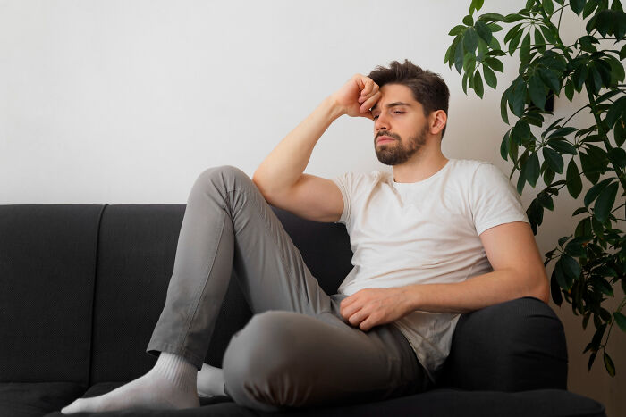 Man in a white shirt and gray pants sitting on a couch, looking thoughtful, next to a green plant.