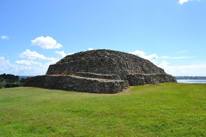 Ancient man-made stone structure on green field under blue sky near a body of water, showcasing historic construction techniques.