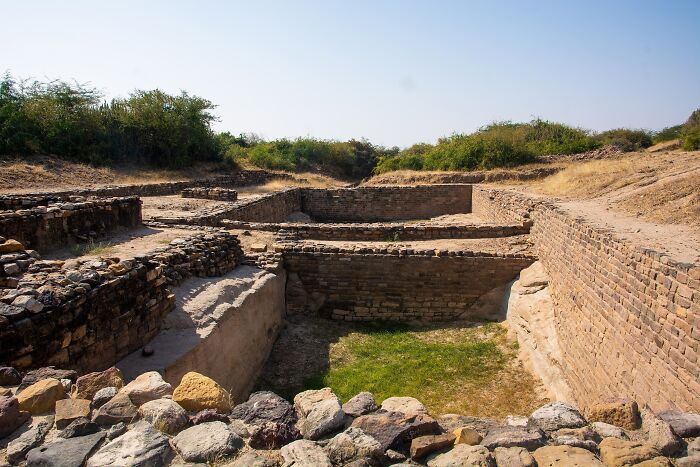Ancient man-made stone structure ruins surrounded by dry grass and bushes under a clear blue sky.