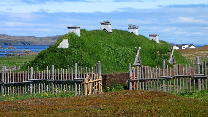 Ancient man-made structures with grass-covered roofs near a coastal area surrounded by wooden fences under a blue sky.