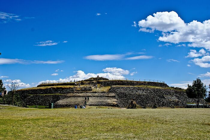 Ancient man-made stone structure under a blue sky with scattered clouds, surrounded by grass and trees.