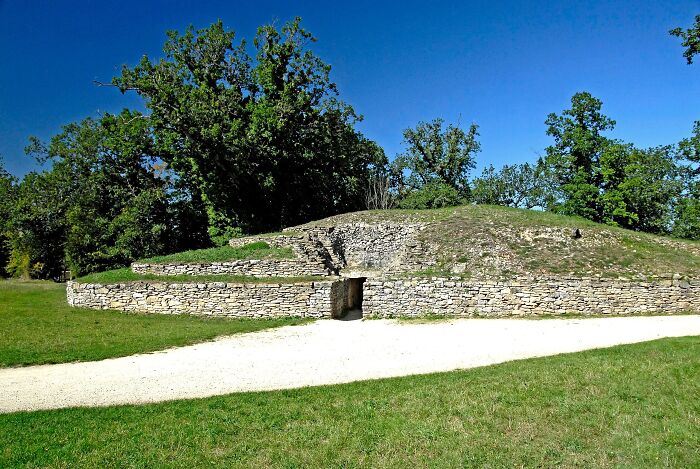 Ancient man-made stone mound structure surrounded by grass and trees under a clear blue sky.