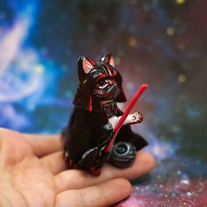 Hand holding a pocket-sized animal figurine in a black cloak with a red lightsaber against a cosmic background.