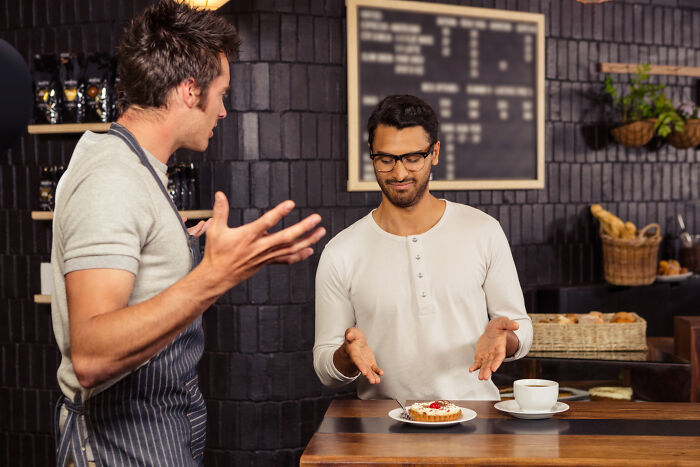 Barista gesturing at a customer with a tart and coffee, illustrating masterful insulting skills in a cafe setting.