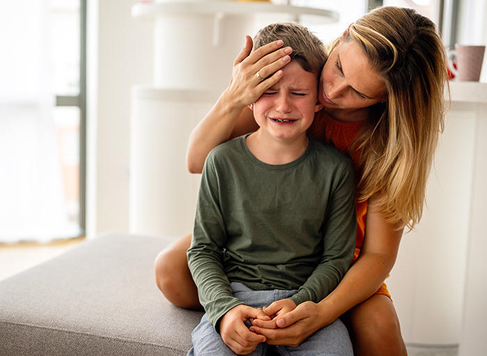 A concerned mom comforts her crying child, sitting on a couch in a bright room. A concerned mom comforts her crying child, sitting on a couch in a bright room.