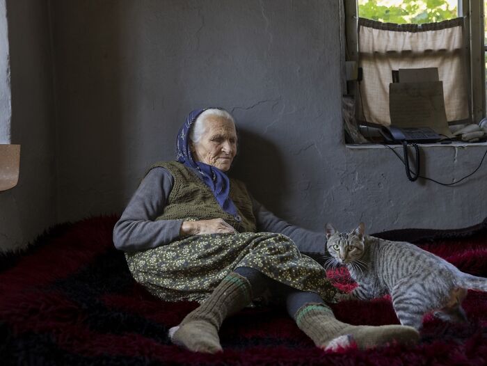 An elderly woman in a headscarf and woolen socks sits on a rug with a cat, illustrating the vulnerability of aging.