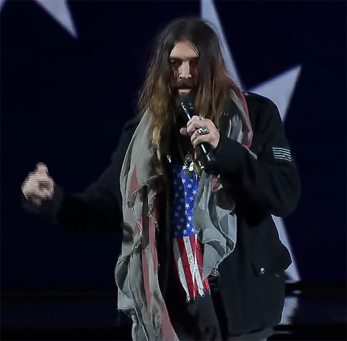Man with long hair on stage, holding a microphone, wearing a scarf with American flag details. Man with long hair on stage, holding a microphone, wearing a scarf with American flag details.