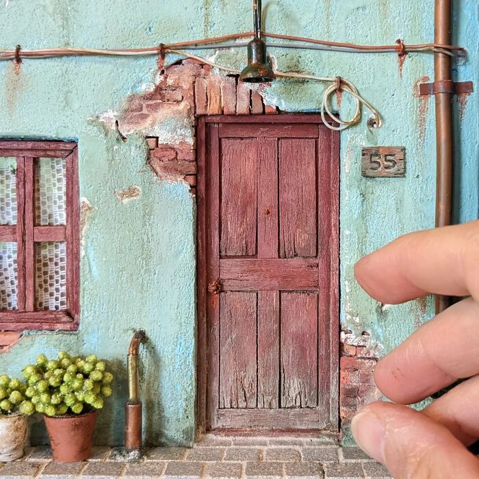 A hand reaches towards a rustic miniature door marked "55" on a textured wall, showcasing detailed tiny worlds.