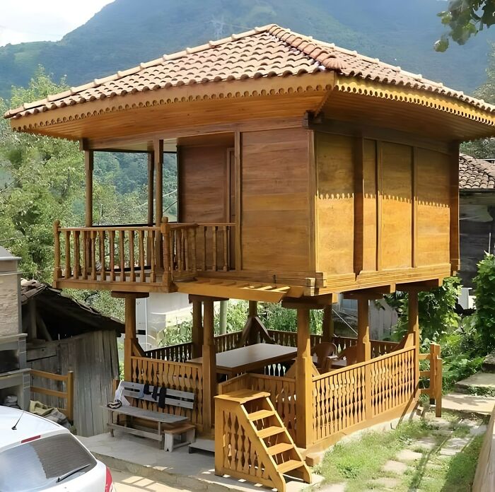 Two-story wooden house with intricate woodworking details, elevated design, and a tiled roof in a natural outdoor setting.
