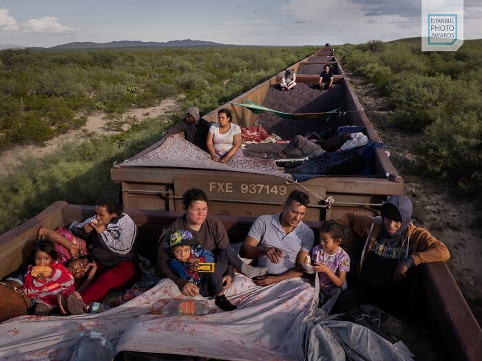 People traveling on an open train carriage through a rural landscape, highlighting social themes from Istanbul Photo Awards 2025.