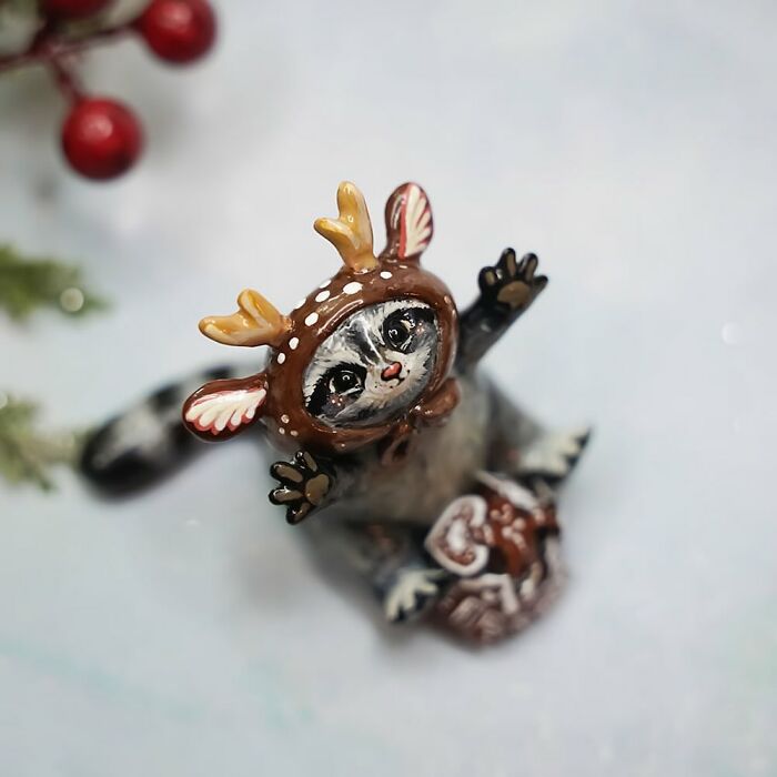 Pocket-sized animal figurine by Vallavica, depicting a raccoon in a cute deer costume on a snowy background.