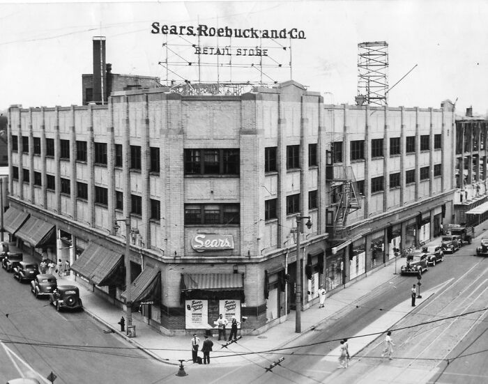 Historic Sears Roebuck store building with vintage cars and pedestrians, showcasing life in America from 100 years ago.