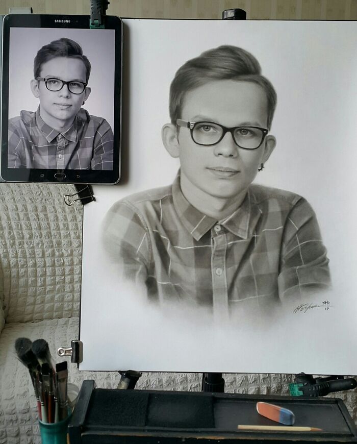 Hyper-realistic pencil portrait on easel with reference photo of a person wearing glasses and a plaid shirt.