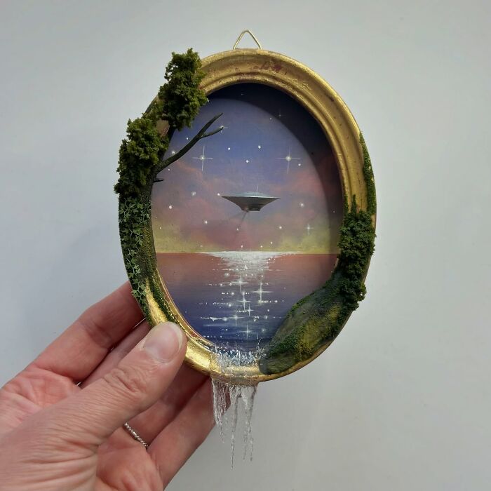Hand holding a diorama with a UFO over water, encased in an oval frame with tree and moss details.