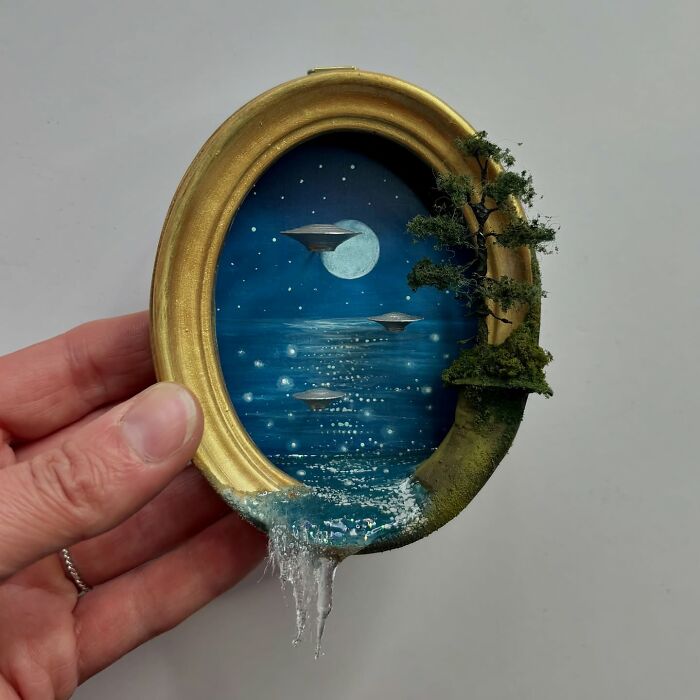 Miniature diorama with UFO scene, crafted by Caroline Dewison, featuring a golden oval frame and ocean landscape.