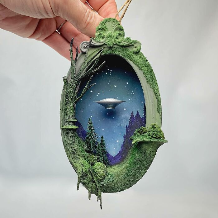 Alien diorama by Caroline Dewison depicting a UFO over a forest, encased in an oval frame with greenery.