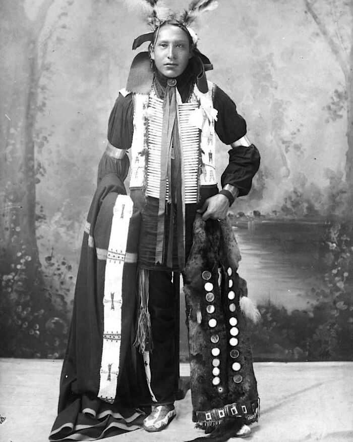 Native American man in traditional dress holding a decorated fur robe, representing real lives from a century ago.