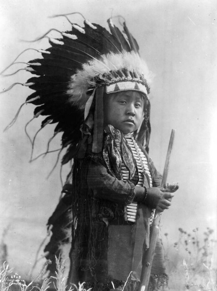 Young Native American boy wearing a traditional feathered headdress and holding a wooden staff in a historic black and white photo.