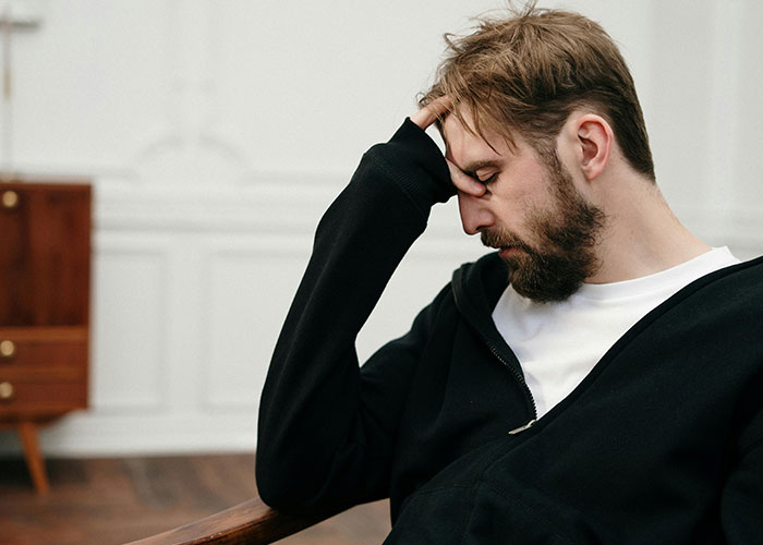 Man in a black hoodie sitting, appearing frustrated and thoughtful, hand on forehead in a home setting. Man in a black hoodie sitting, appearing frustrated and thoughtful, hand on forehead in a home setting.