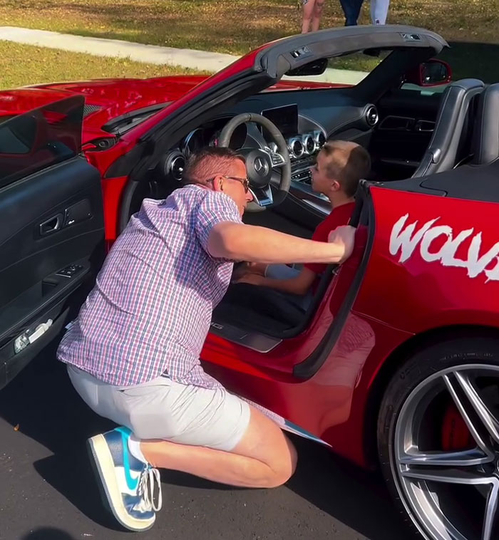 Boy sits in a red sports car as a person kneels beside, smiling during a birthday surprise from local car enthusiasts. Boy sits in a red sports car as a person kneels beside, smiling during a birthday surprise from local car enthusiasts.