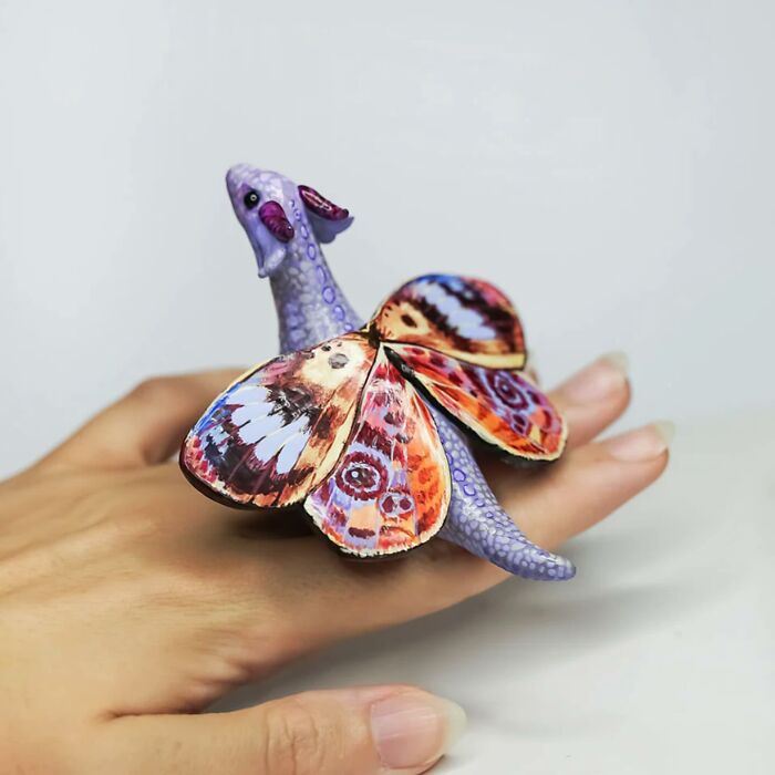 Pocket-sized animal figurine by Vallavica, featuring a purple creature with colorful wings resting on a hand.