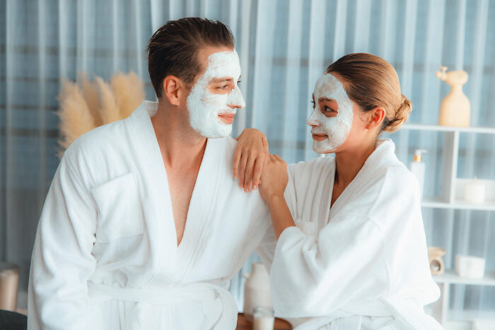 Couple in spa robes, smiling with face masks on, sharing a moment together.