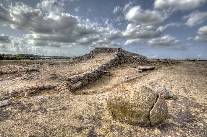 Ancient man-made stone structure with c*****d large boulder in a dry landscape under dramatic cloudy sky.