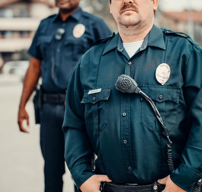 Two police officers in uniform standing outdoors, focus on one officer's badge and radio, representing authority and control. Two police officers in uniform standing outdoors, focus on one officer's badge and radio, representing authority and control.