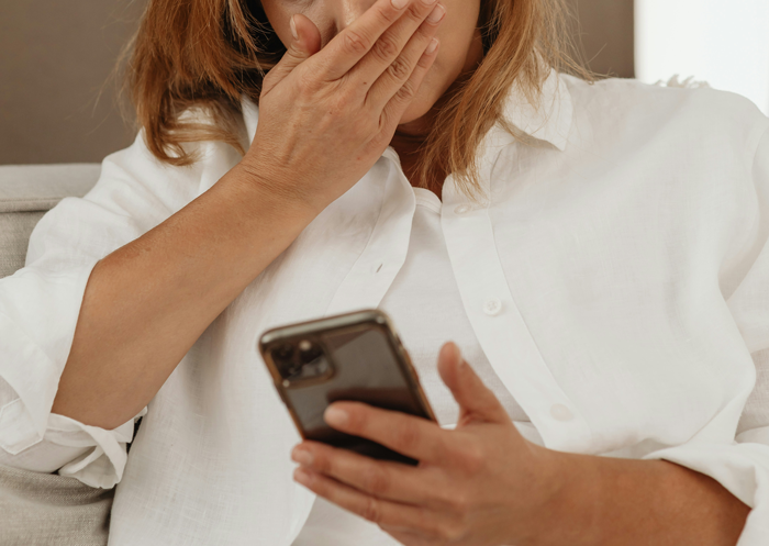 A woman in a white shirt looks shocked at her phone, covering her mouth. A woman in a white shirt looks shocked at her phone, covering her mouth.