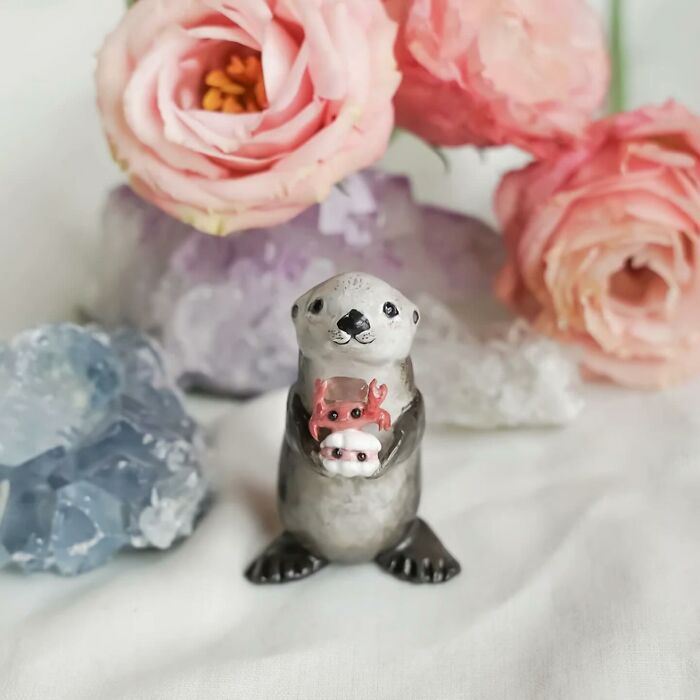 Pocket-sized animal figurine of an otter holding a small pink crab, surrounded by pink flowers and crystals.