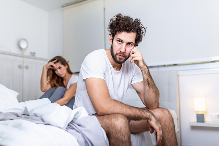 Man looking pensive sitting on edge of bed while woman in background looks upset, exploring why guys slept with married woman.