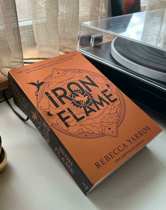 Book titled "Iron Flame" by Rebecca Yarros resting on a table near a turntable, with curtains in the background.