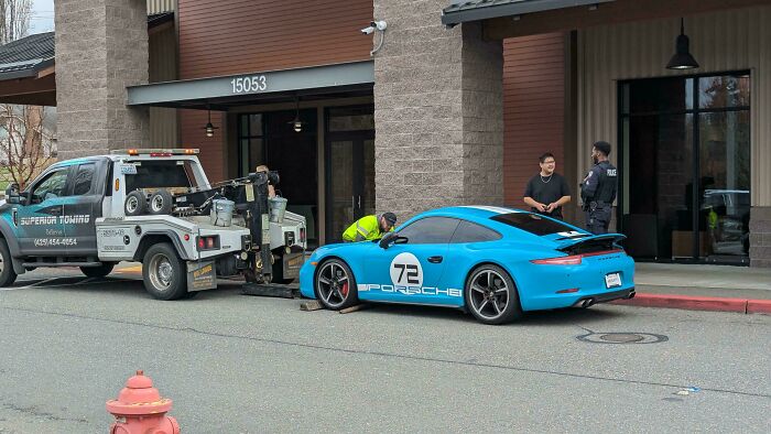 Tow truck preparing to tow a bright blue Porsche outside a building, highlighting lack of self-awareness.