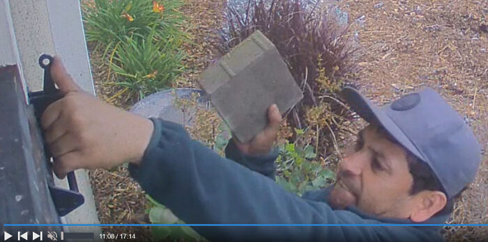 Man lacking self-awareness uses a brick to adjust a window latch, wearing a cap outside.