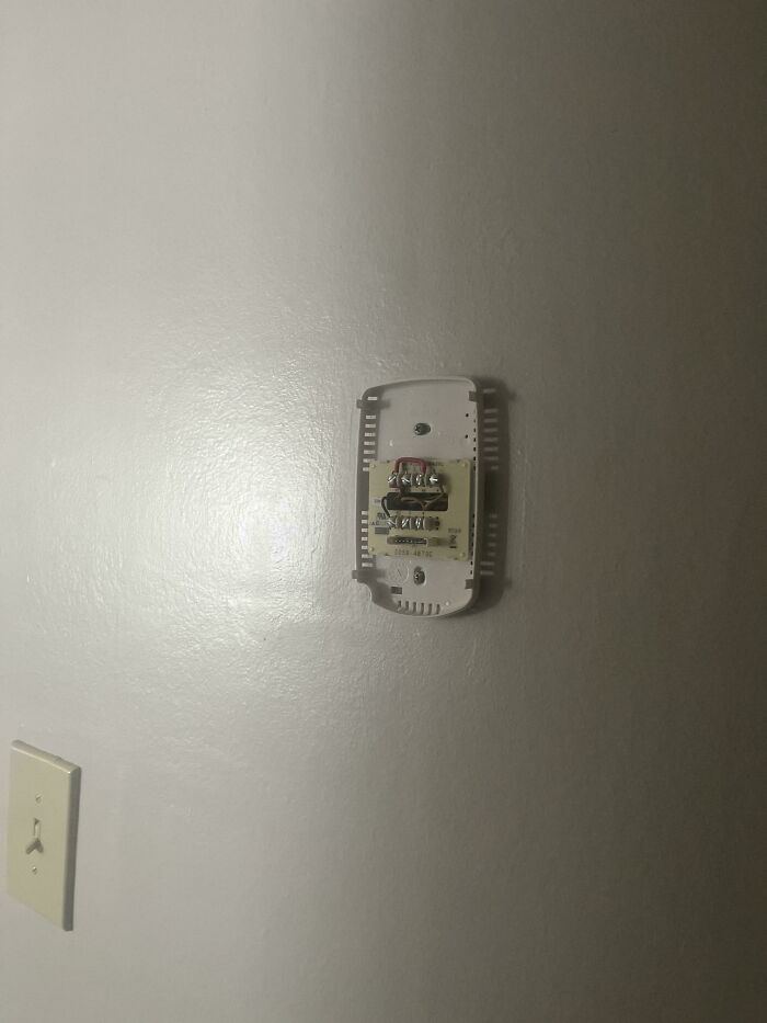 Thermostat without cover on a plain wall, highlighting lack of self-awareness in home maintenance.