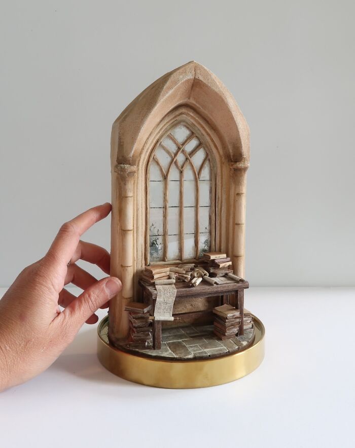 A hand holds a miniature scene by Tamar Cohen, featuring a tiny arched window and desk with books.