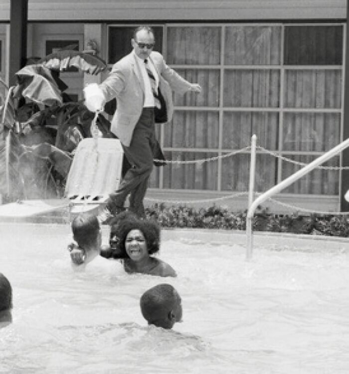 A man steps into a hotel pool fully clothed, expressing anger and surprising swimmers around him.