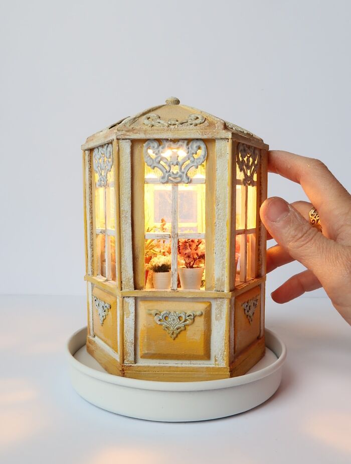 A hand touches a lit miniature greenhouse by Tamar Cohen, showcasing intricate tiny worlds.