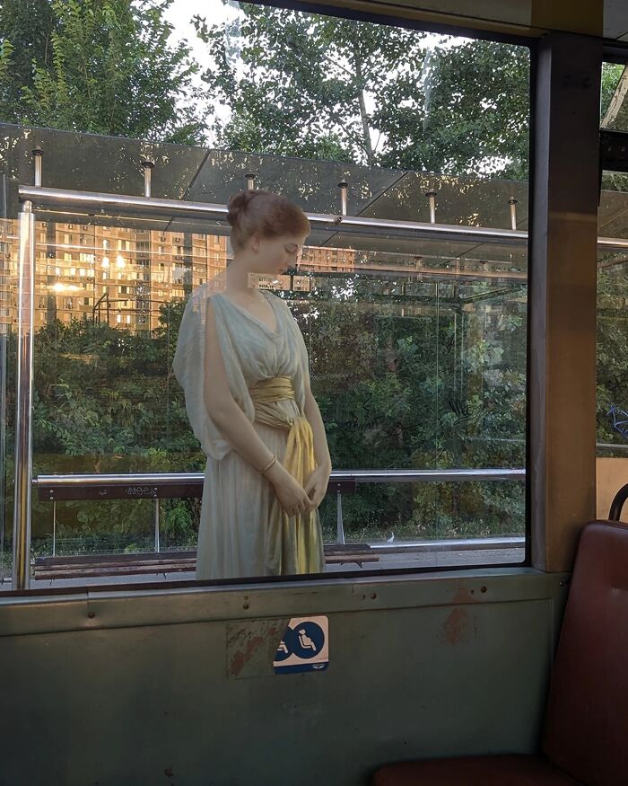 Classical figure in modern setting, standing outside a bus window with trees and buildings in the background.
