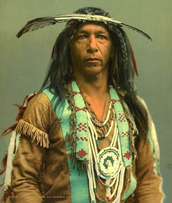 Native American man wearing traditional Ojibwa clothing and feathered headdress in a historic portrait photo.