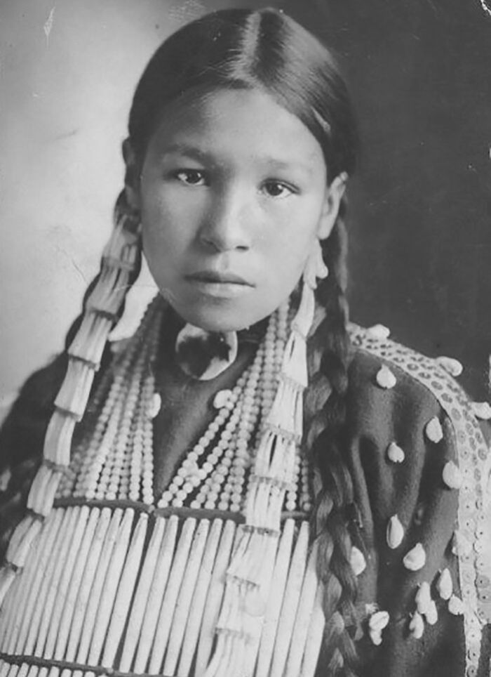Young Native American girl in traditional attire with beads and braids, a haunting photo revealing real lives a century ago.