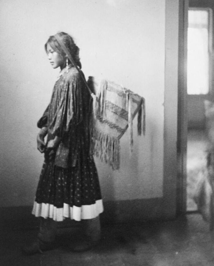 Native American woman in traditional clothing carrying woven blanket in a vintage black and white photo.