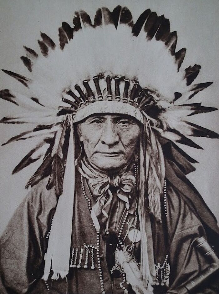Native American elder in traditional feathered headdress and ceremonial attire in a haunting historic portrait.