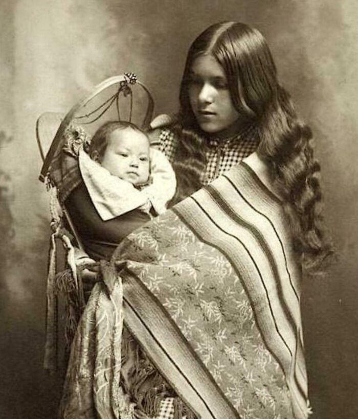 Young Native American woman holding baby wrapped in patterned blankets in haunting early 20th century photo.