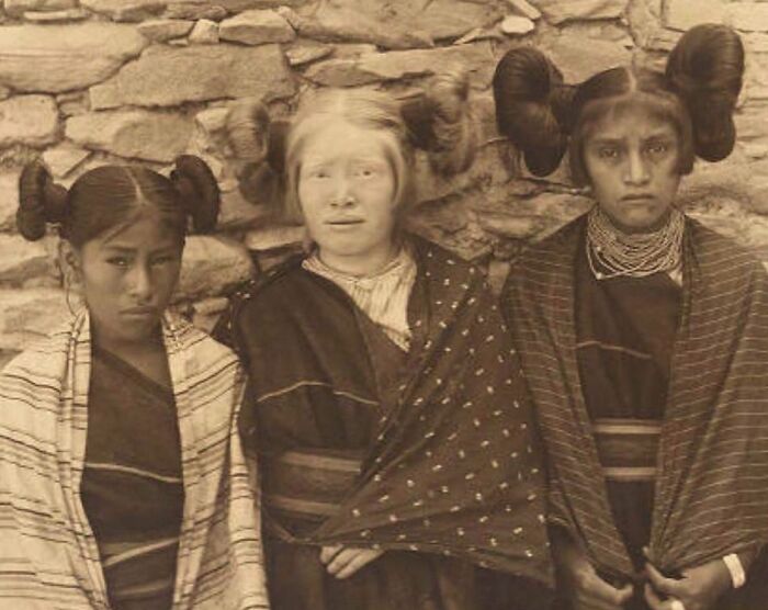 Three Native American girls with traditional hairstyles and clothing posing against a stone wall in a historic photo.